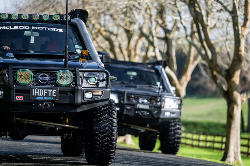 Two modified Nissan Patrol off-road touring vehicles driving through rural New Zealand with aftermarket 4x4 accessories